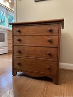 Front angled view of a wooden four-drawer chest with round knob handles on wooden floor near a kitchen stove and window.