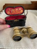 Opened black hinged case with red interior and gold embossed design showing opera glasses removed and placed nearby on beige surface