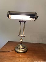 Brass metal piano desk lamp lit from front view showing cylindrical shade and round base.