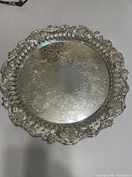 Front view of a round silver tray showing intricate embossed floral pattern on the surface and ornate cut-out floral border.