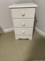 Front view of small white wooden night stand or dresser with three drawers, each with round white knob handles, paint shows some wear.