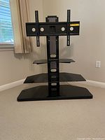 Front view of the black glass TV stand showing adjustable mounting bracket and three shelves.
