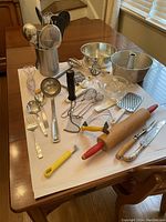Various kitchen utensils arranged on a wooden table including ladles, whisks, peelers, graters, rolling pin, egg beater, strainers, and forks.