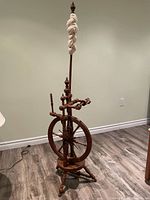 Full upright view of the antique wooden spinning wheel with distaff holding spun fiber, positioned on a wood floor.