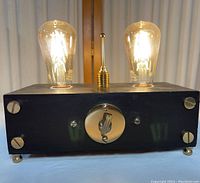 Front view showing black rectangular wooden base with two lit Edison bulbs and central gold toggle switch with circular plate.