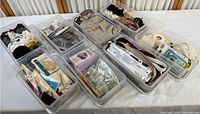 Seven plastic containers holding various sewing and craft supplies such as ribbons, trims, elastic bands, threads, needles, and sewing tools, arranged in a row on a surface.