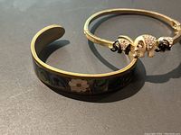 Two cuff bracelets shown on surface in sunlight. One bracelet has abalone shell floral design, the other has elephant motifs with rhinestones and enamel.