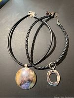 Photo showing two modern necklaces: one with a large round multicolored metal pendant on a smooth black cord, the other with a small irregular oval silver-tone pendant on a braided black cord with adjustable chain and clasp.