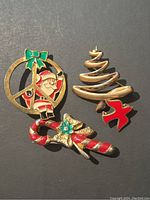 Four vintage Christmas-themed jewelry brooches on dark background showing Santa in circle with green bow, spiral Christmas tree, candy cane with gold bow, and red bird.