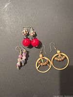 Three pairs of vintage dangle earrings displayed on a black surface: red bead clip-ons, purple bead dangles, beige hoop dangles with brown beads.