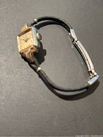 Front angled view of the watch showing the gold filled case, black fabric strap and metal clasp with visible dial and crown.