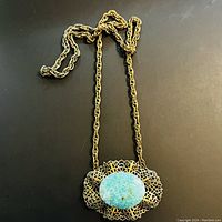 Full view of natural stone large pendant with delicate filigree metal ornate setting on a gold-tone chain necklace laid flat on surface.