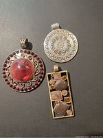 Three pendants displayed on black background: large round pendant with red cabochon and purple stalks, round silver-tone engraved pendant, rectangular multistone gold-tone pendant.