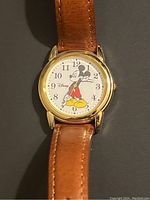 Front view of vintage Disney Mickey Mouse watch showing the round gold-tone case, white dial with Mickey Mouse illustration, black numbers, and brown leather strap.