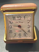 Front and slightly angled view of vintage Seth Thomas mini portable alarm clock in open brown leatherette folding case, showing cream clock face with Arabic numerals, black hour and minute hands, red alarm hand and the brand name signature.