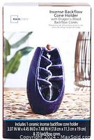 Photo showing ceramic incense waterfall backflow cone holder and box on a woven surface with greenery background, box label reads Mainstays Incense Backflow Cone Holder with Dragon's Blood Backflow Cones