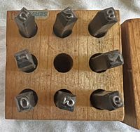Top-down photo of the wooden case with steel number stamps inserted into slots, showing digits 0,1,2,3,5,6,8,9 present and missing #4.