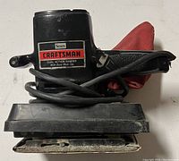 Side profile photo showing the black body of the Craftsman sander with red dust pick-up bag and attached power cord coiled around the handle.
