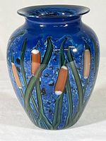 Front view of signed hand-blown glass vase displaying cattail and green stem design on deep blue background
