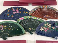Five decorative folding hand fans with floral designs laid out with original red boxes