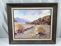 Full view of framed painting showing desert scene with mountains, rocky cliffs, and shrubs under sky with textured brush strokes.