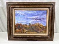 Full view of the framed oil landscape painting showing a cloudy sky over a path with autumn-colored bushes and rocks. Dark wood frame with beige inner border.