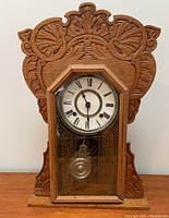 Front view of baby grandfather clock showing carved wooden case, octagonal bezel with Roman numerals, and etched glass door closed.
