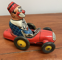 Side angled view of the vintage tin clown in a red racing car toy showing its colorful paint and four wheels.