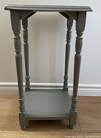 Grey wooden side table with turned legs and lower shelf shown standing on wooden floor.