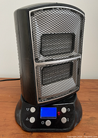 Front view of the black and gray ceramic heater showing dual heating elements behind mesh grille, control buttons, and blue LED screen on base.