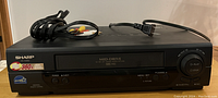 Front view of Sharp VC-A850 VCR player showing front controls, tape slot and branding with cables on top.