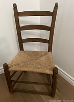 Front angle view of a small antique wooden ladderback chair showing woven natural fiber seat and three horizontal slats on the backrest.