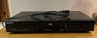 Front view of the black Sony DVD Player model DVP-NS710H showing front control buttons, display panel, DVD and CD logos, and attached power cord.