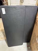 Two black folding tables stacked against a wall, showing their soft textured tops and 4ft length.