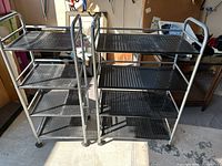 Two metal rolling utility carts side by side, showing their size difference and design with perforated shelves and caster wheels.