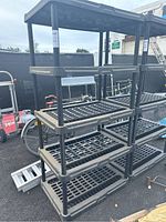 Two black 5-tier plastic shelving units outdoors on asphalt, showing their five grid-style shelves and tubular supports.