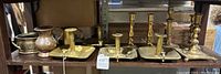 Shelf view of assorted antique brass candle holders with vessels including chamberstick types and taller candlesticks, showing patina and some wear.