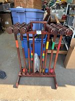 Wooden croquet set with six mallets, balls, wickets, and stand stored indoors with background items visible.