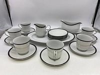 Front view of 6 white ceramic tea cups with black rim, each paired with a saucer; a creamer, gravy boat, and large platter placed behind
