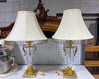Pair of glass and brass table lamps with white bell-shaped fabric shades trimmed with beaded fringe sitting on a table.