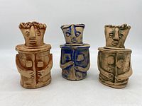 Front view of three handmade lidded clay art pottery pieces shaped as stylized human figures in terracotta, blue, and tan clays