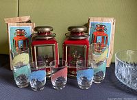 Photo showing 7 glasses with colorful feather designs arranged in front of 2 red oil lamps with brass tops and original boxes.