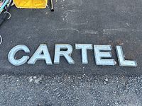 Full set of metal light up letters spelling 'CARTEL' laid out on gravel and asphalt surface.