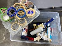 Photo of multiple rolls of painters tape in plastic container, along with assorted painting tools such as paint rollers, brushes, and trays.