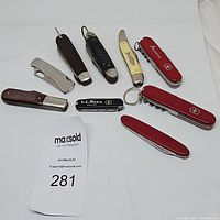 Nine folding pocket and camping knives arranged on white surface including multiple Victorinox and L.L.Bean branded items.