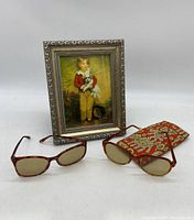 Two pairs of sunglasses, decorative fabric holder, and picture frame arranged on white surface