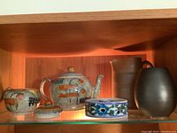 Six artisan pottery pieces displayed on a glass shelf inside a wooden cabinet, showing teapot, sugar bowl, decorative box, and two pitchers.