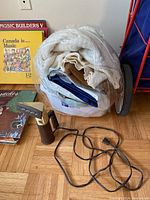 Clear plastic-wrapped bedding and clothing bundle with knits in neutral tones along with brown Franzus steamer with power cord on wooden floor and assorted books in background.