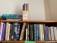 Row of hardcover books and novels in varied subjects arranged upright on a bookshelf with a cardboard box labeled 371 and Sellers Books