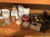 Multiple candles and black metal candle holders displayed on wooden surface, showing variety in shape, size, and decorative style.